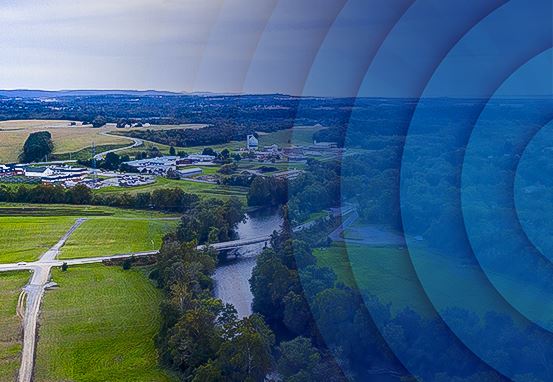 Aerial view of a river flowing through green fields and trees with rural buildings in the background under a blue sky.