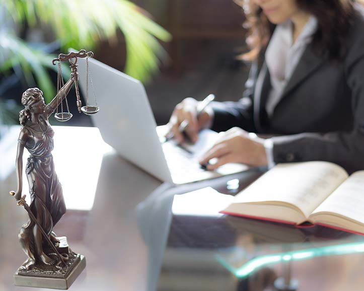 Bronze Lady Justice statue on desk with open book and a woman in business attire working on a laptop in the background.