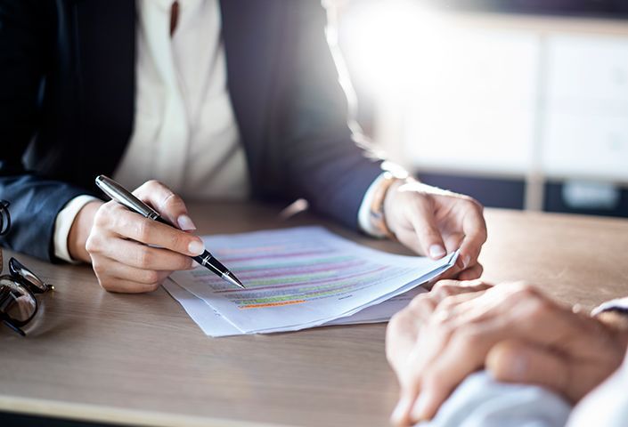 A person in a suit points at colorful highlighted text on a document with a pen. Another person listens intently in a sunlit office setting.