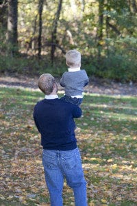 A person in a dark sweater and jeans holds a young child on their shoulders while walking through a sunlit, wooded area with fallen leaves.