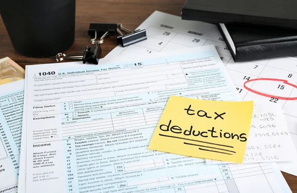 An IRS Form 1040 sits on a desk with a calendar and a yellow sticky note that reads tax deductions in handwritten black ink.