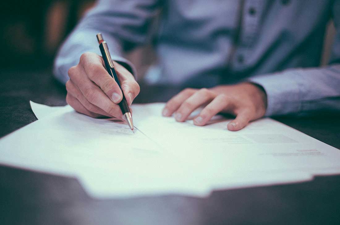 A close-up shot of a person’s hands writing on a stack of papers with a pen, resting on a dark wooden desk.