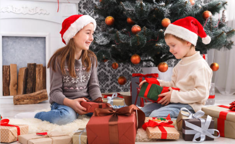 Two children wearing Santa hats unwrapping Christmas gifts in a living room.
