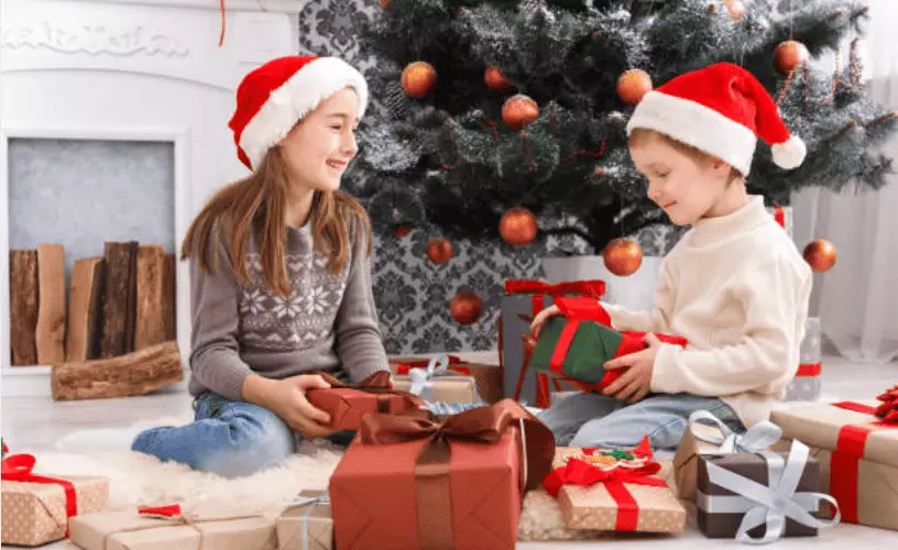 Two children wearing Santa hats unwrapping Christmas gifts in a living room.