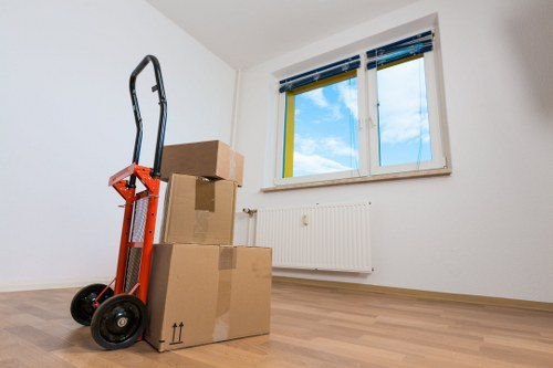 A red hand truck loaded with cardboard boxes stands in the middle of a bright, empty room with white walls and a large window