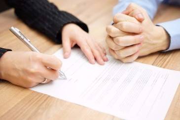 A close-up of two people's hands over a document on a wooden desk. One person is writing with a pen while the other has their hands clasped.