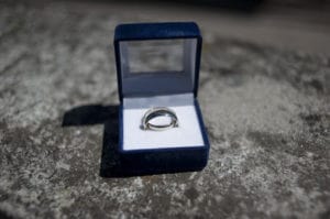 An open dark blue jewelry box sits on a textured surface, showcasing a silver-colored ring or bracelet nestled inside on a white cushion.